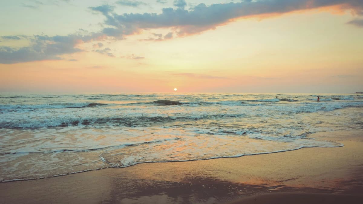 Waves wash onto a sandy beach at sunset, with the sun low on the horizon and a few swimmers far out in the sea
