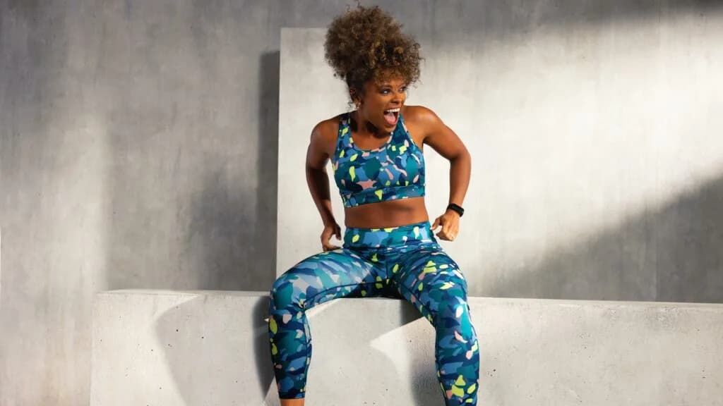 Woman with curly hair in a blue patterned sports bra and leggings sits on a concrete block, laughing with her mouth open