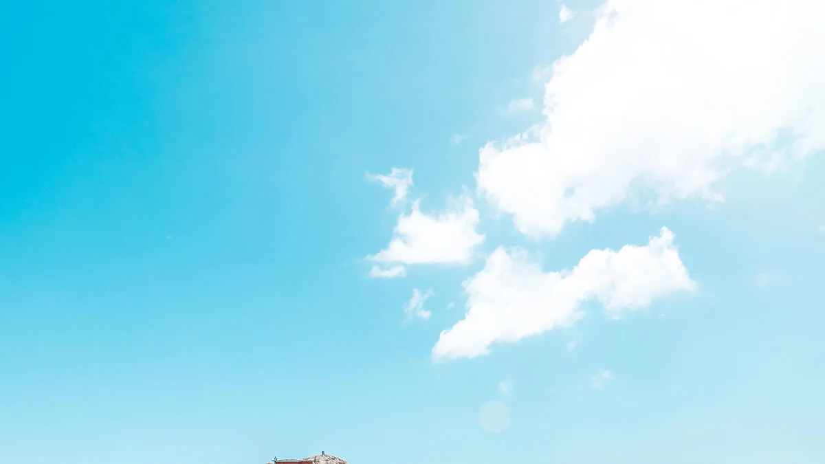 Bright blue sky with white clouds above the thatched roof of a small red building at the bottom edge
