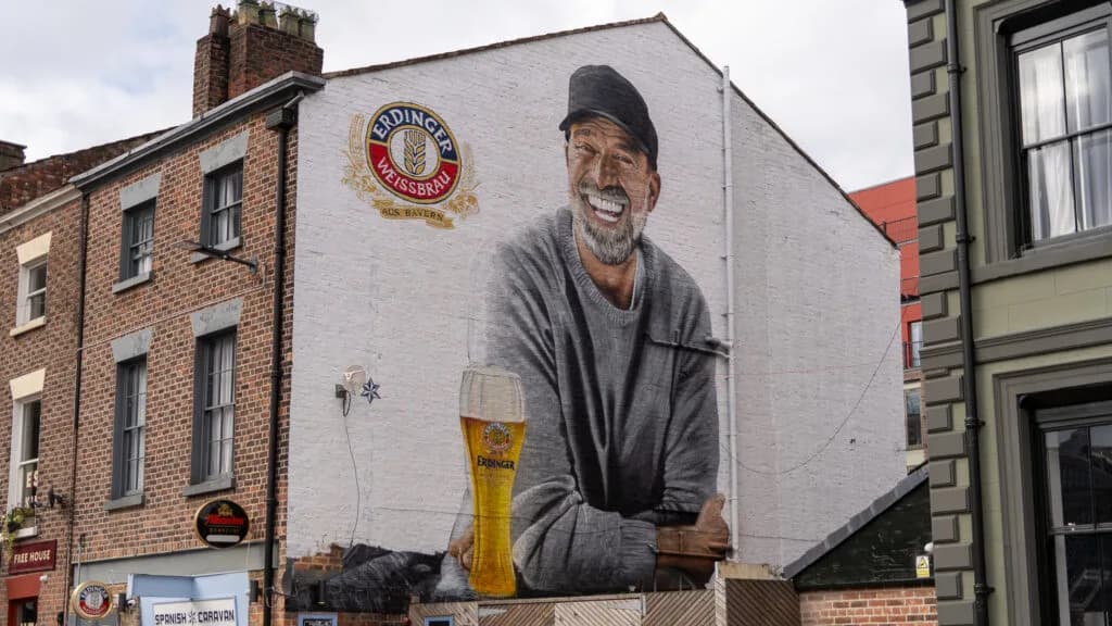 Street mural of a grinning bearded man in a cap leaning beside a giant glass of ERDINGER wheat beer on a building wall