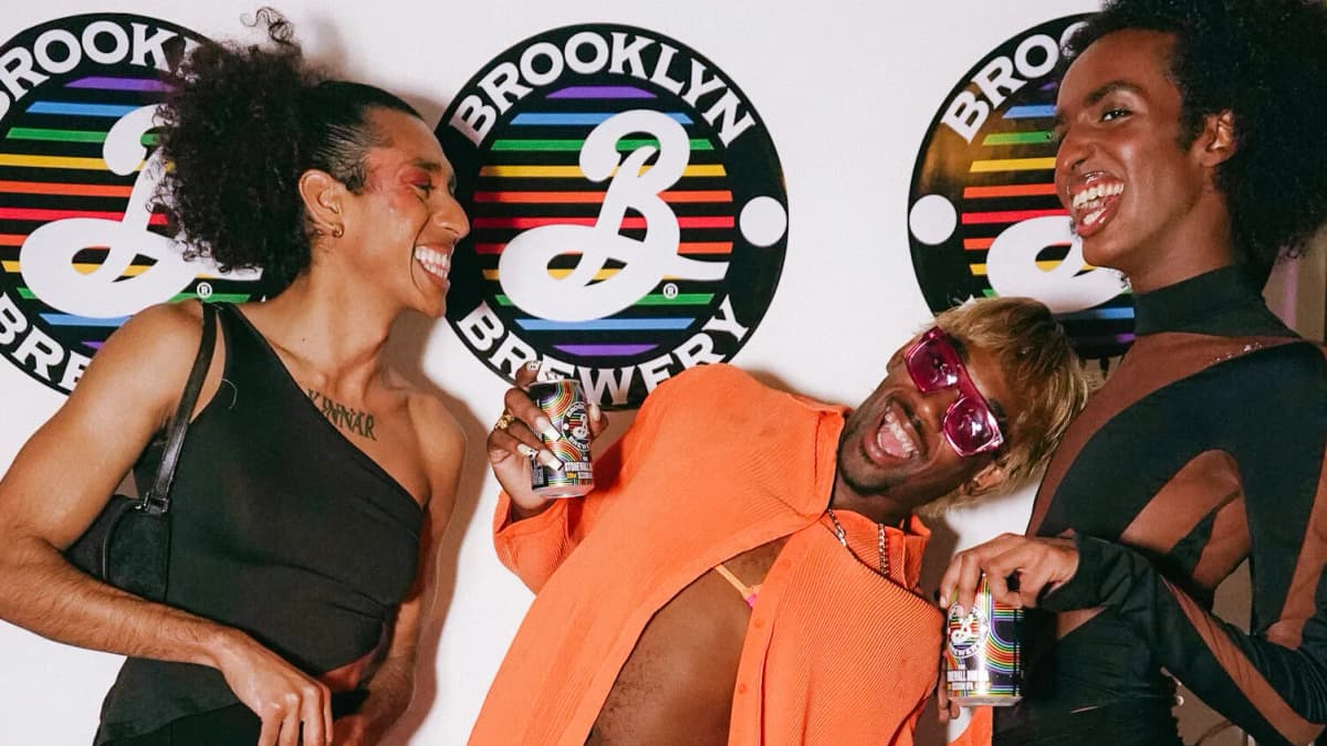 Three laughing people, two holding Brooklyn Brewery cans, pose before a rainbow "BROOKLYN BREWERY" logo backdrop