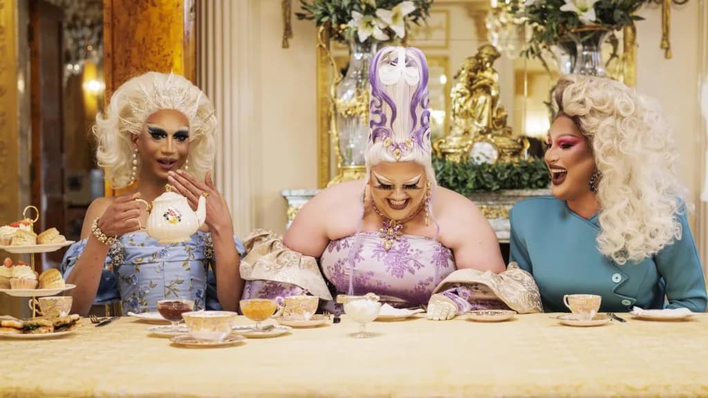 Three drag queens in big wigs and gowns laugh over afternoon tea, teapot and cakes, in an ornate gilded room