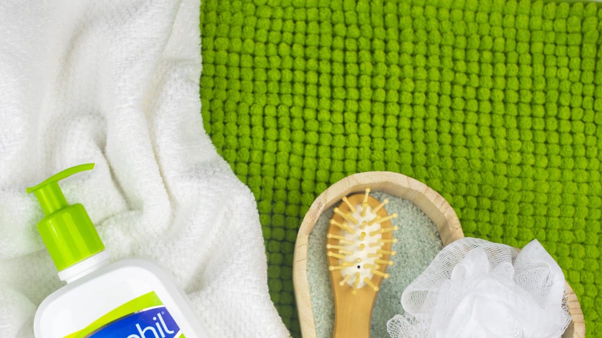 Cetaphil pump bottle on a white towel beside a wooden bowl holding a hairbrush and bath sponge on a green mat