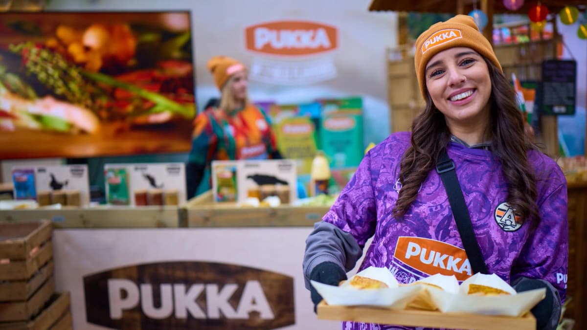 Smiling woman in a "PUKKA" beanie and purple shirt holds a tray of pie samples at a Pukka stall with a "PUKKA" banner