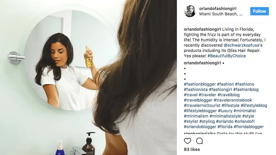 Woman in a white T-shirt sprays oil from a gold bottle onto her long dark hair, reflected in a round bathroom mirror