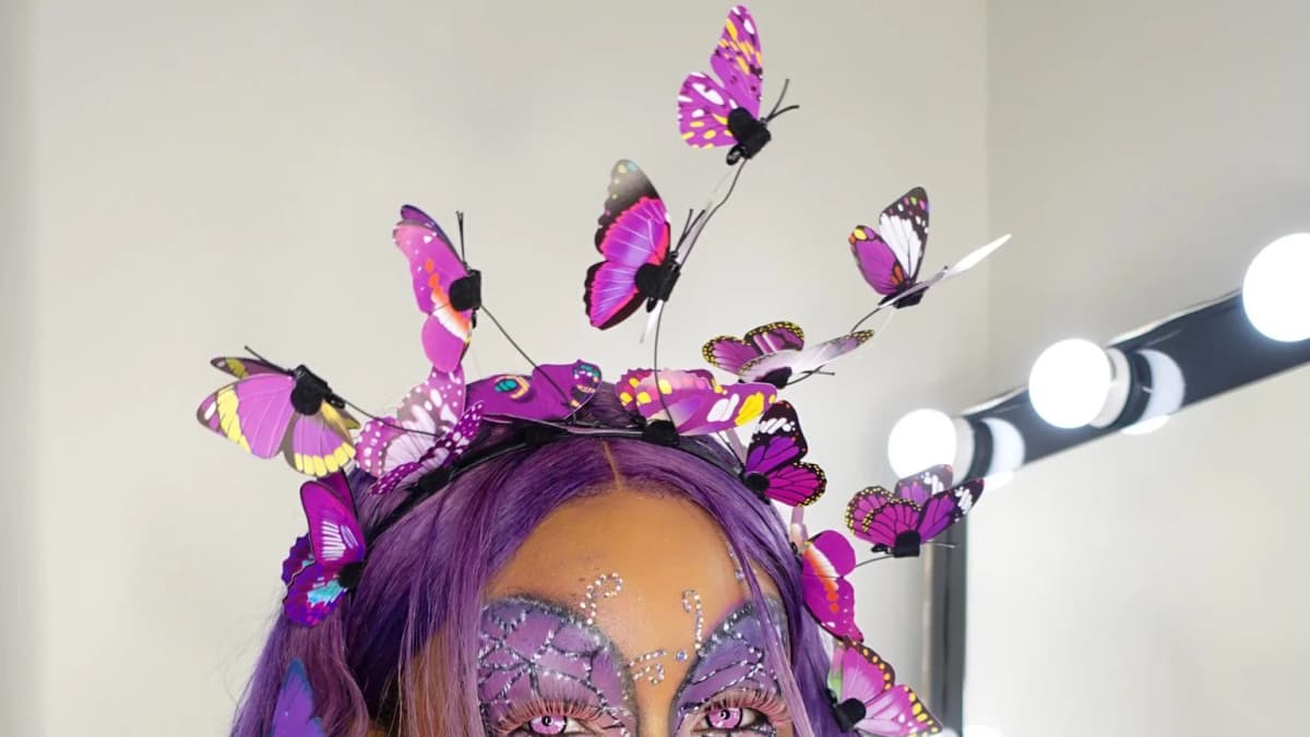 Person with purple hair, a crown of purple butterflies and jeweled butterfly face makeup beside a lit vanity mirror