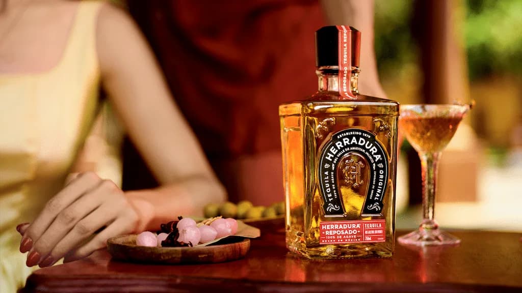 Herradura "Reposado" tequila bottle and a cocktail glass on a wooden table beside snacks, with two people seated behind