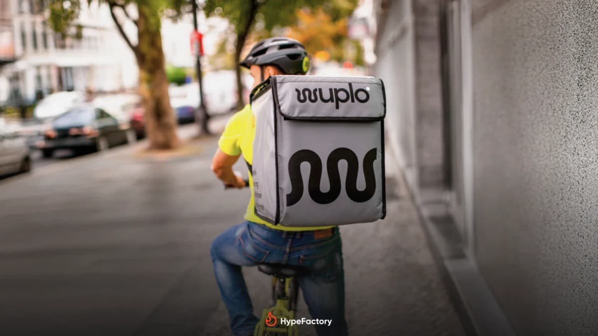 Cyclist in a helmet and yellow shirt seen from behind on a city street, carrying a grey "wuplo" delivery backpack