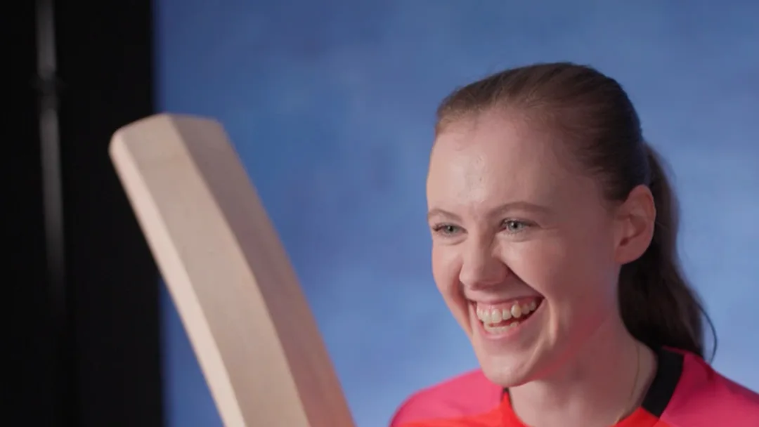 Smiling woman with a ponytail in a pink sports shirt holds up a cricket bat in front of a blue studio backdrop