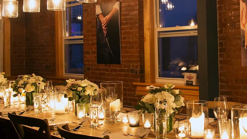 Candlelit dinner table with white flowers and glassware in a brick-walled room beside a poster of hands holding a knife