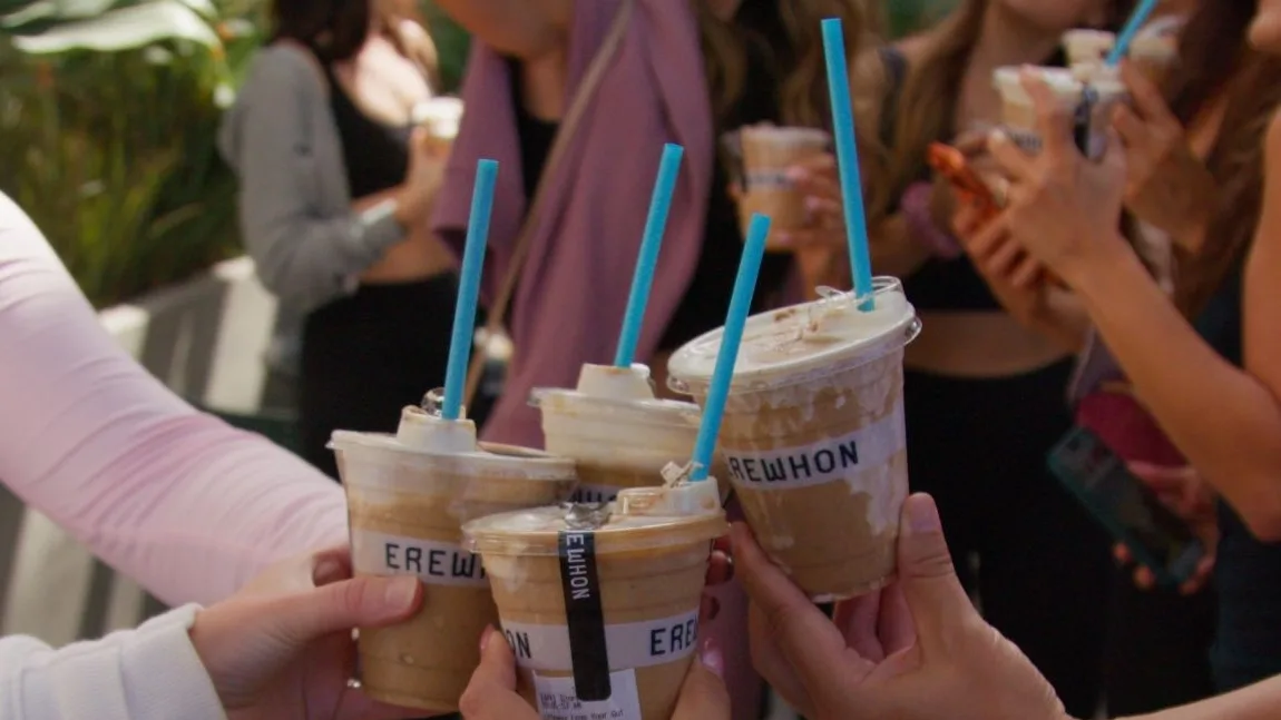 Hands toast four iced smoothies with blue straws and whipped tops in labeled cups, as a group of women stands behind them
