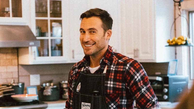 Smiling man in a plaid flannel shirt and dark denim apron stands in a bright white kitchen with pots on the stove