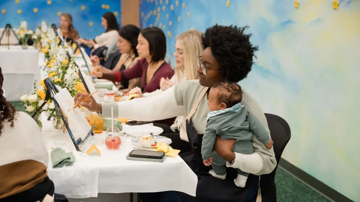 Woman holding a baby paints on a small easel beside other women at a long table with flowers and a sky mural