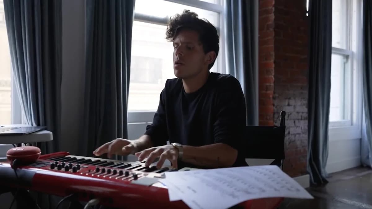 Man in a black sweater plays a red keyboard with sheet music beside him in a loft with tall windows and brick walls