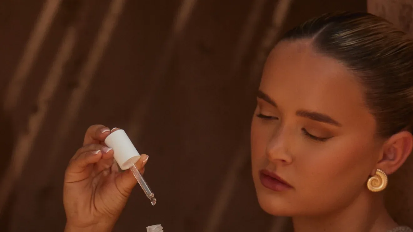 Woman with slicked-back hair and gold spiral earring holds a white dropper over a small bottle, eyes closed, in warm light