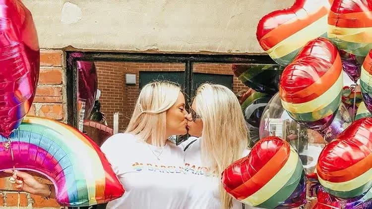 Two blonde women in white Barclays T-shirts kiss among rainbow heart and rainbow-shaped balloons by a brick wall