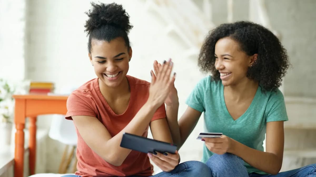 Two smiling young women with curly hair high-five at home, one holding a tablet and the other a bank card