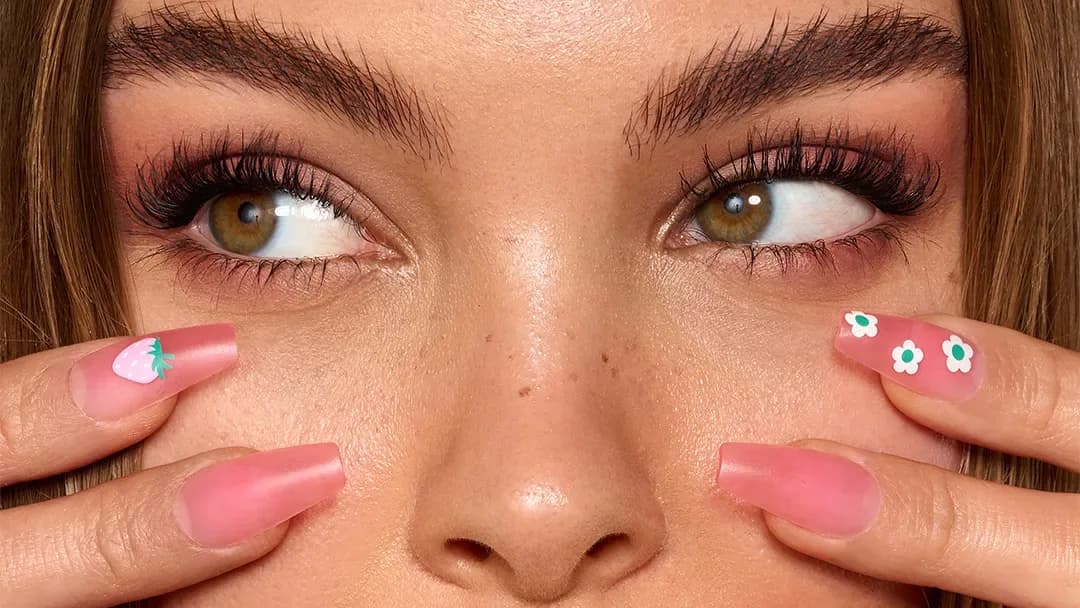 Close-up of a woman with long lashes glancing sideways, fingertips showing pink nails with strawberry and daisy art