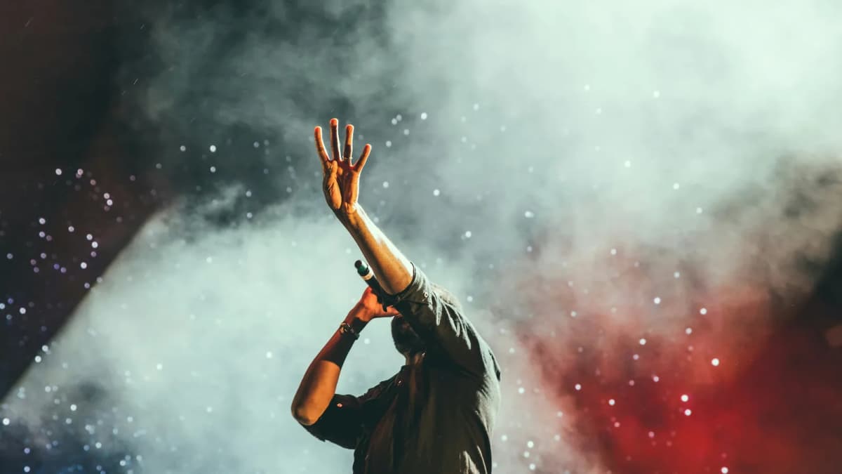 Performer in a dark shirt raises one hand while singing into a microphone on stage, with smoke and red lights behind