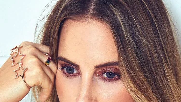Close-up of a woman with long brown hair and blue eyes, hand near her temple, wearing star-shaped rings