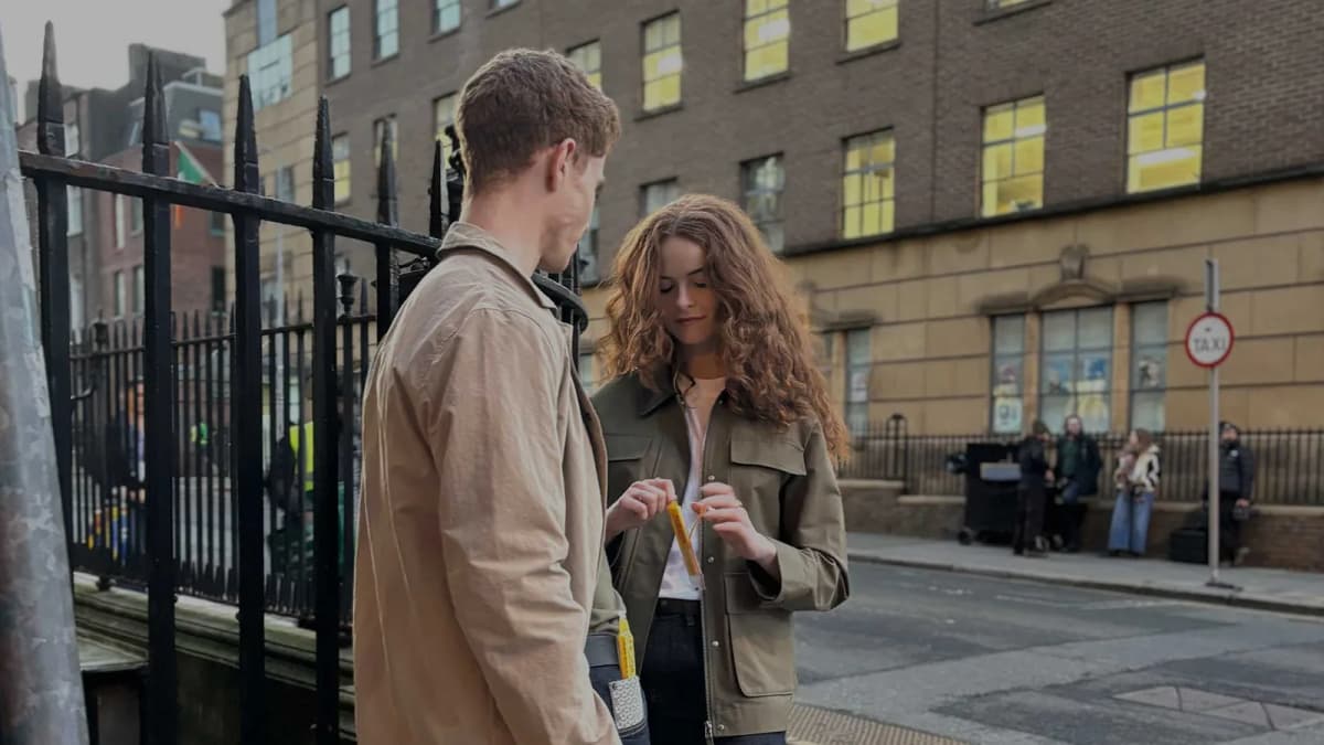 Curly-haired woman opens a yellow cheese snack stick beside a man in a tan jacket on a city street by iron railings