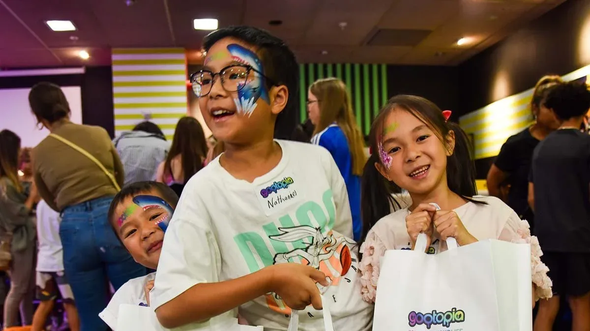 Three smiling children with face paint hold white "gootopia" gift bags in a crowded indoor venue