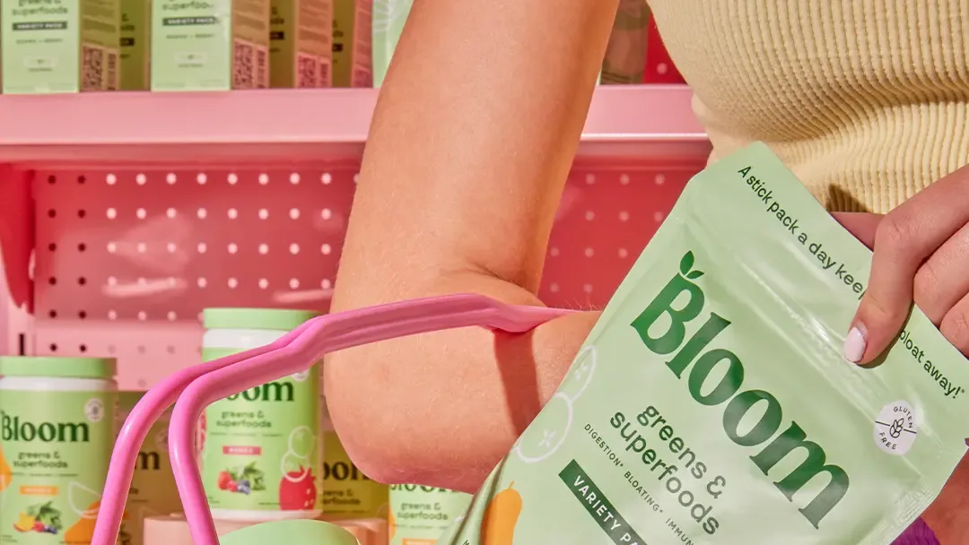 Person with pink basket on arm holds a Bloom "greens & superfoods" pouch by pink shelves stocked with Bloom canisters