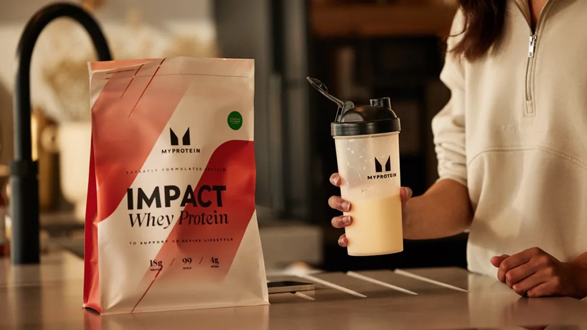 Person in a cream zip top holds a MyProtein shaker beside an "IMPACT Whey Protein" pouch on a kitchen counter