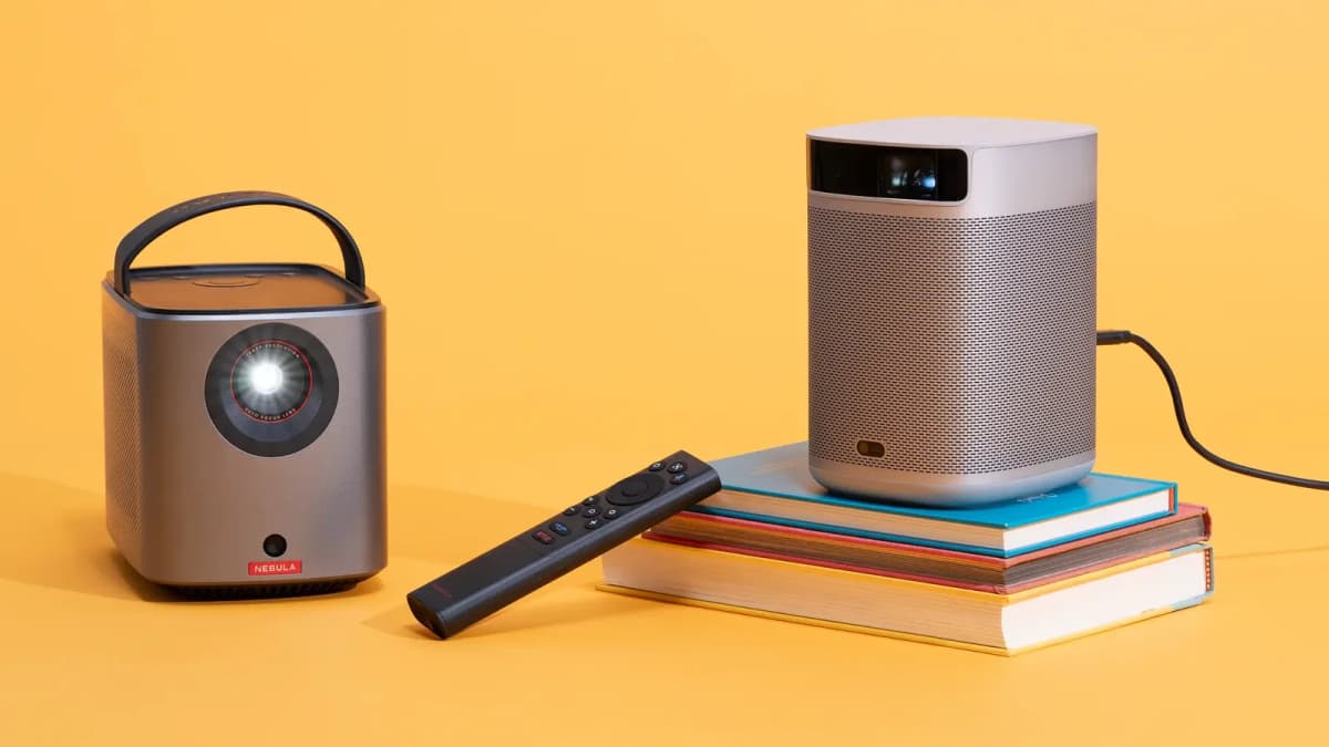 Two portable projectors on a yellow backdrop: one with a handle and lit lens, one silver on stacked books, and a remote