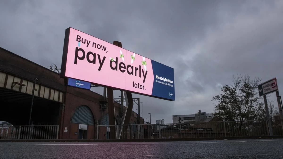 OpenMoney billboard reading "Buy now, pay dearly later." beside brick railway arches under a grey, cloudy sky