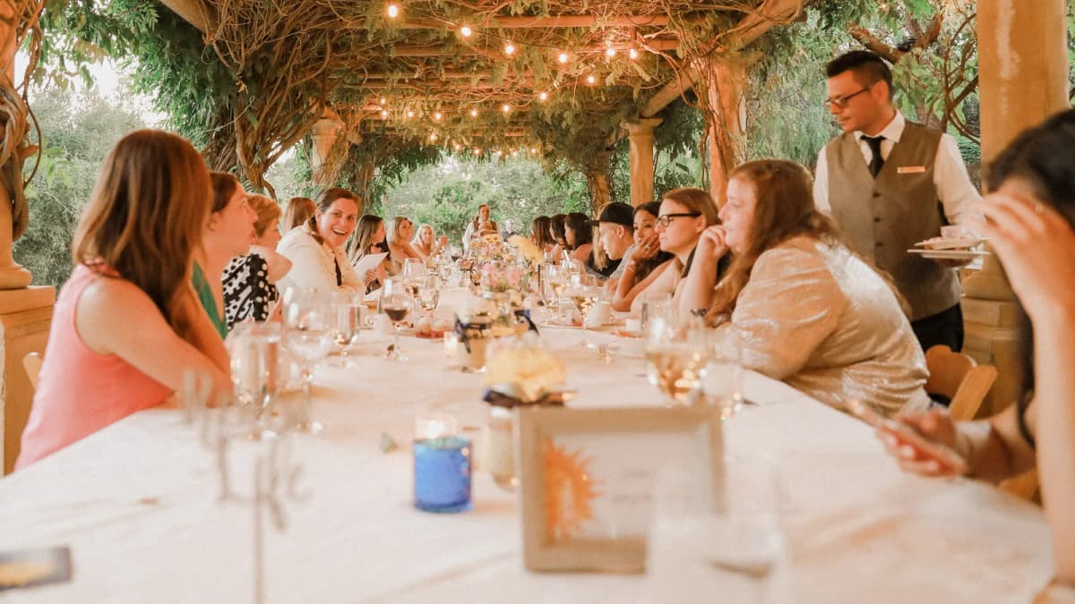 Guests dine at a long white-clothed table under a vine-covered pergola with string lights as a waiter in a vest serves plates