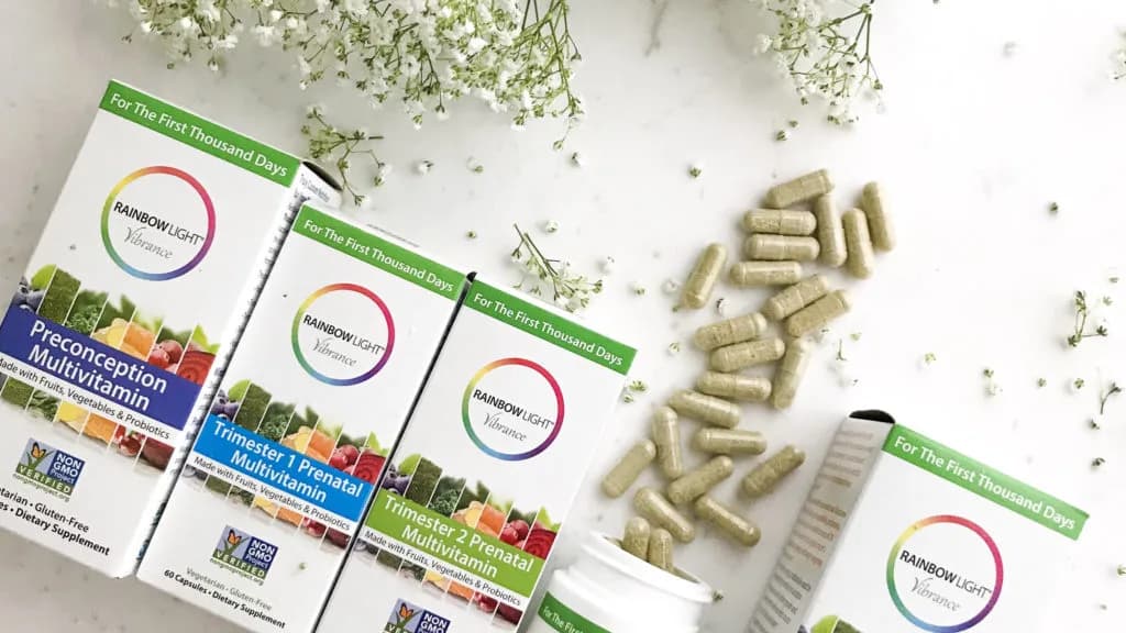 Rainbow Light prenatal multivitamin boxes and green capsules spilling from a bottle beside baby's breath on marble