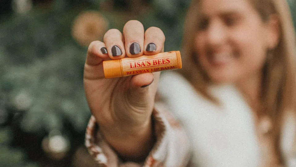 Smiling woman with dark nails holds up an orange Burt's Bees lip balm labeled "LISA'S BEES BEESWAX LIP BALM"