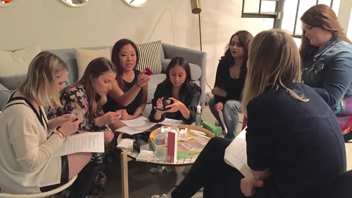 Seven women sit around a coffee table in a lounge, trying makeup products and holding printed sheets of paper