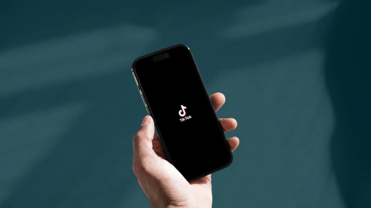 A hand holds a black smartphone showing an app logo on a dark screen against a blurred teal background