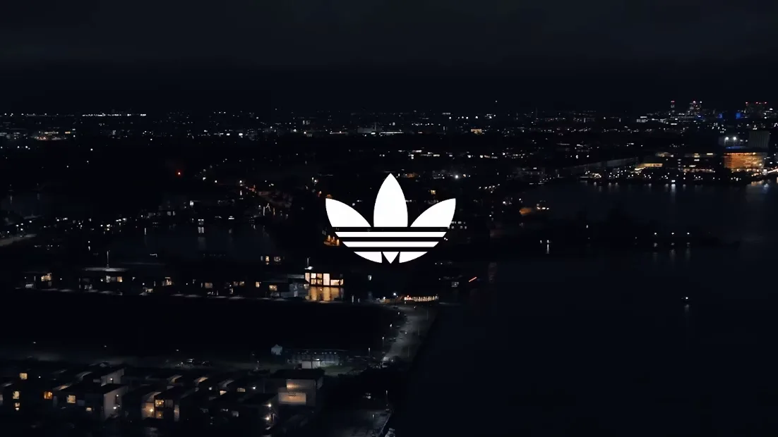 White adidas trefoil logo over an aerial night view of a city with lit buildings along a dark waterfront