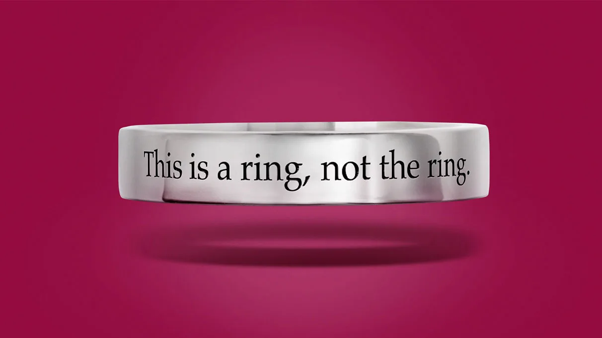 Silver ring band engraved "This is a ring, not the ring." floating above its shadow on a magenta background