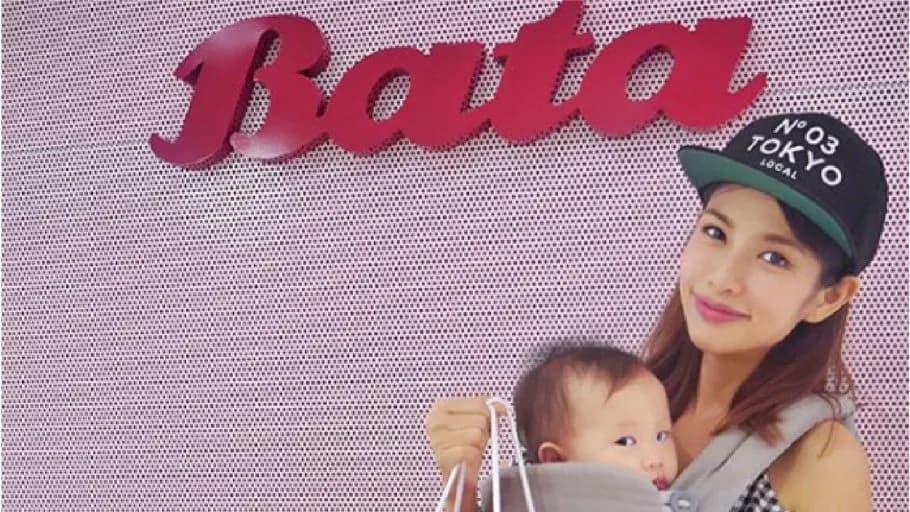 Woman in a "TOKYO" cap carries a baby in a carrier and holds a shopping bag beneath a red Bata logo on a pink wall