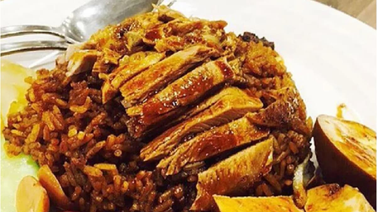 Sliced braised duck on dark soy rice with a braised egg and tofu on a white plate, a metal fork and spoon beside it
