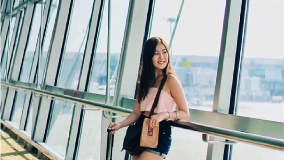 Smiling woman in pink top and denim shorts with a shoulder bag and suitcase, standing by tall windows in an airport terminal