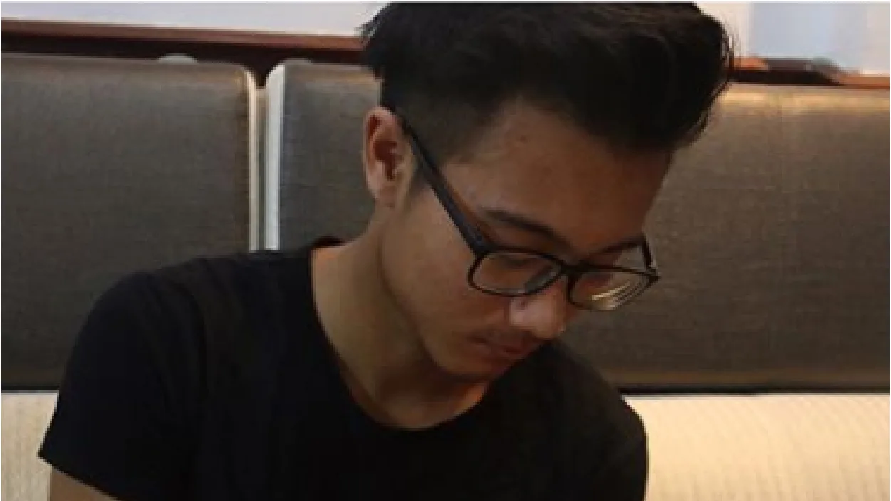Young man with dark hair and glasses, wearing a black T-shirt, looks down while seated on a gray upholstered bench.