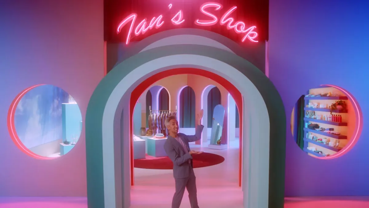 Man in a gray suit gestures from an arched doorway under a pink neon sign reading "Tan's Shop" in a pastel store set