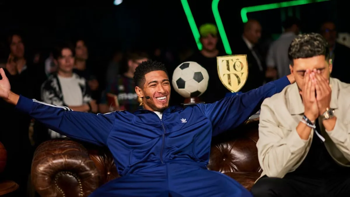 Smiling man in blue tracksuit spreads arms on leather sofa as man nearby covers face; crowd and "TOTY" trophy behind