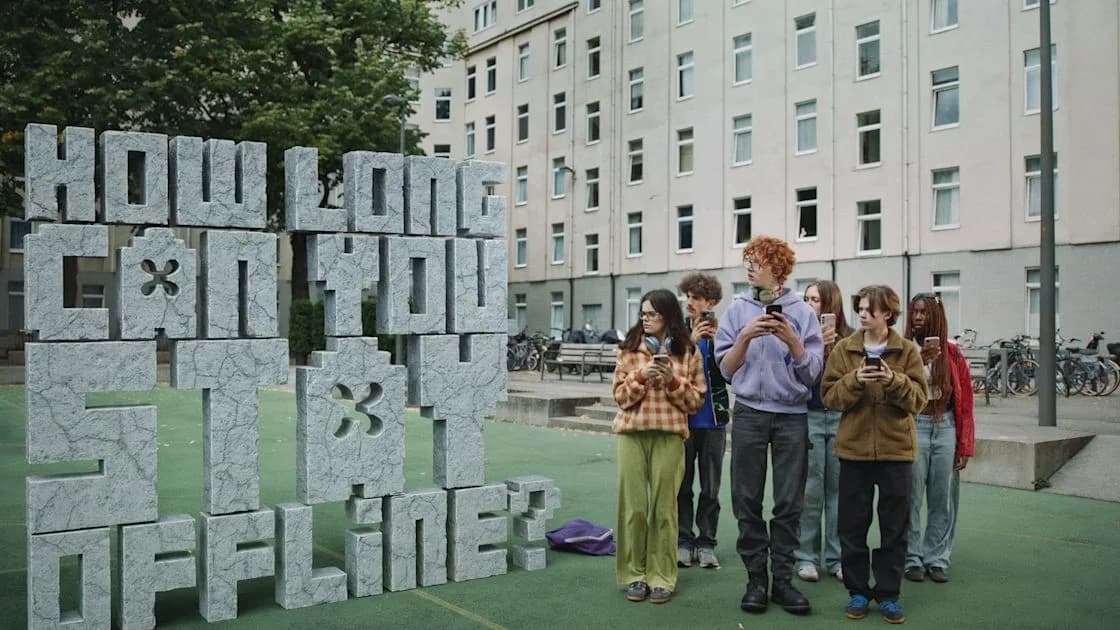 Teens holding phones stand beside a giant stone sculpture reading "HOW LONG CAN YOU STAY OFFLINE?" in a courtyard