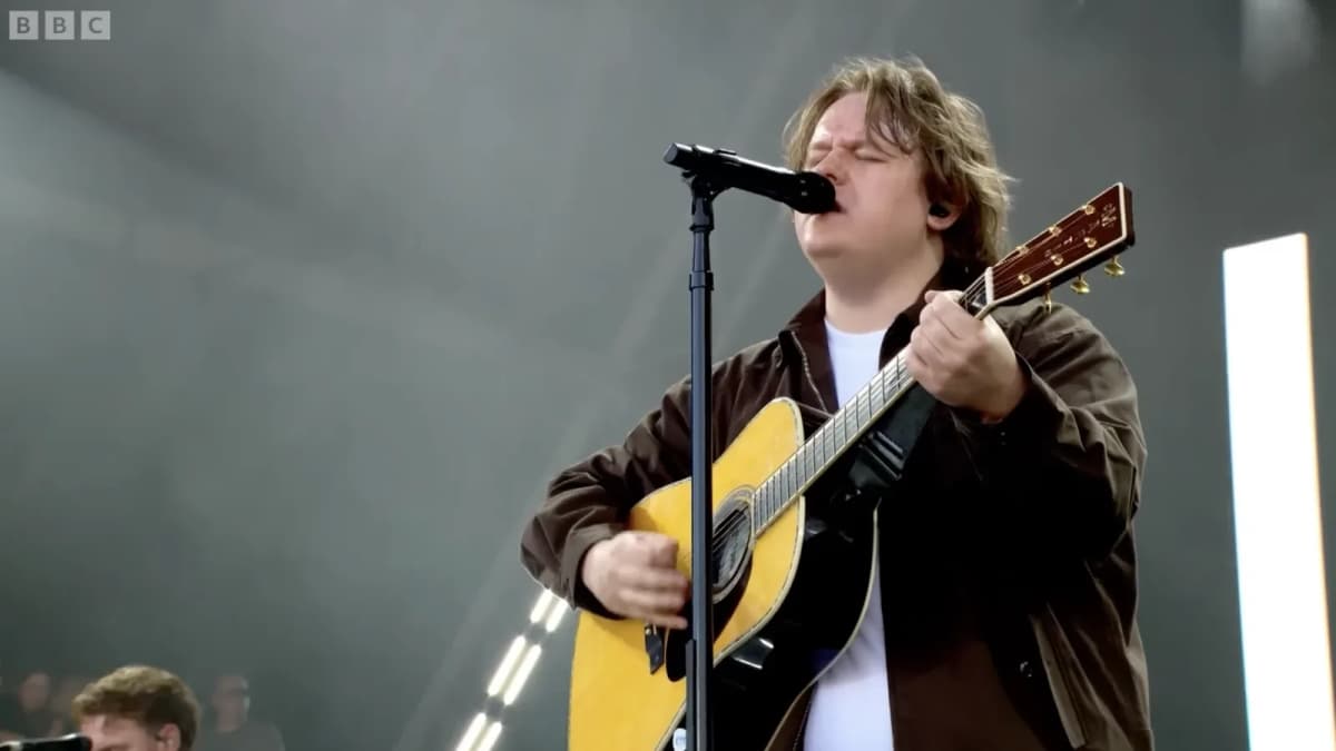 Man with shaggy hair in a brown jacket sings eyes closed into a microphone while playing an acoustic guitar on stage