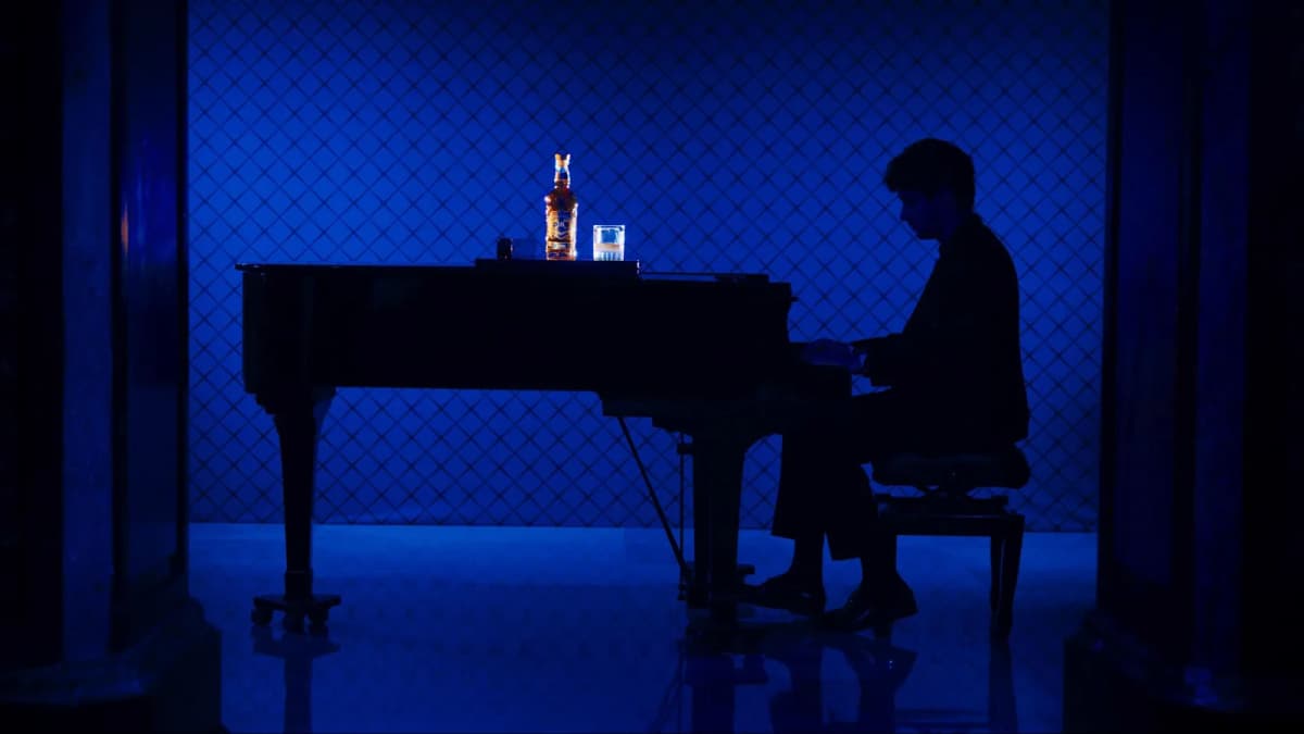 Silhouetted man plays a grand piano in blue light, a lit Chivas Regal bottle and a glass resting on top of the piano
