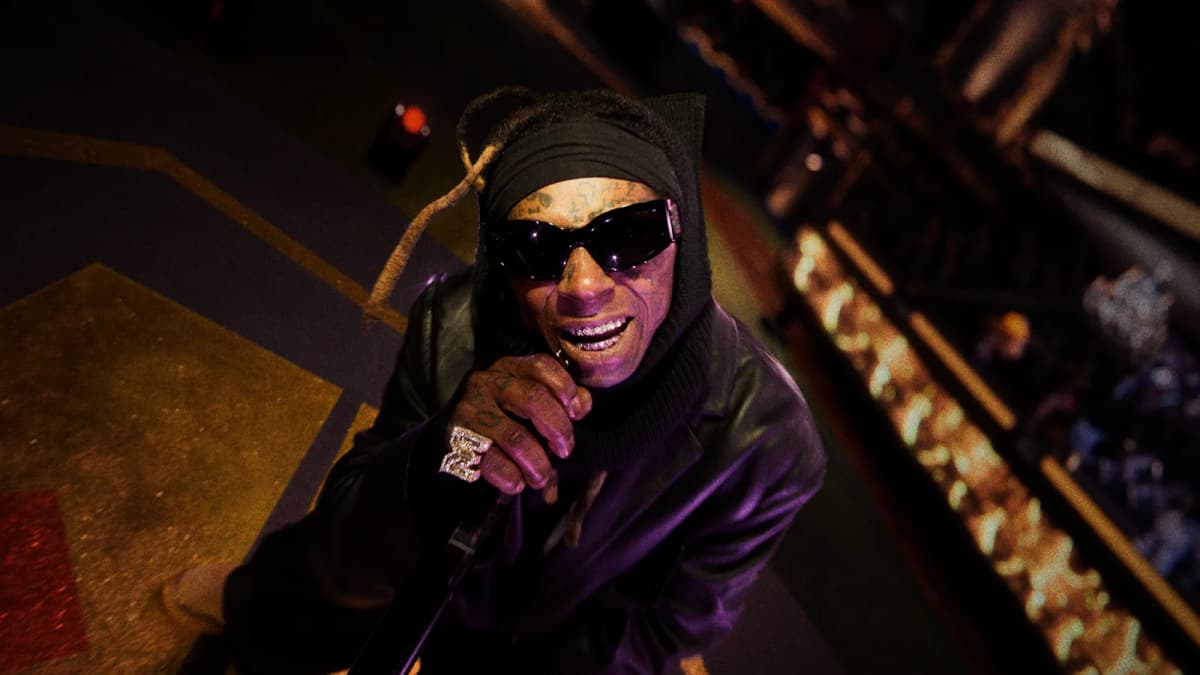 Performer in sunglasses, black head wrap and leather jacket sings into a microphone on a stage lit purple and gold