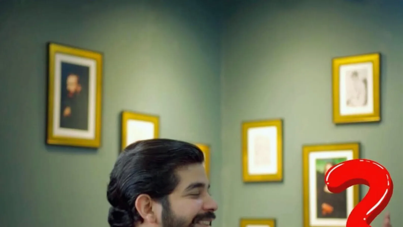 Smiling bearded man with slicked-back hair in a green room with gold-framed pictures, next to a large red question mark