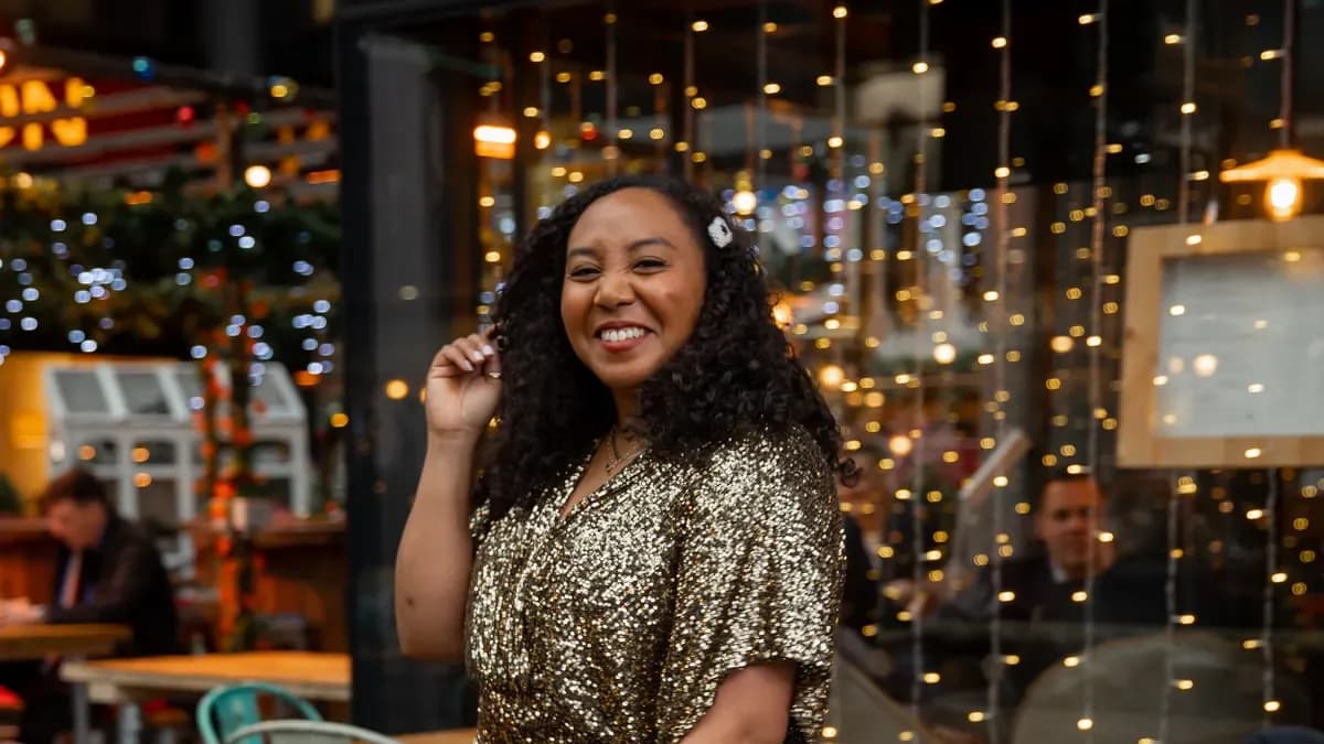 Smiling woman with curly hair in a gold sequin dress poses outside a restaurant window hung with fairy lights