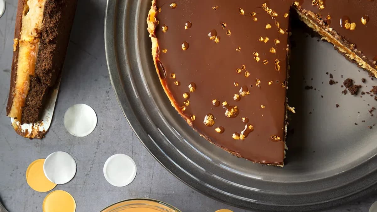 Chocolate-glazed cheesecake with gold flecks in a metal tin, a slice cut out beside it, with gold and silver confetti